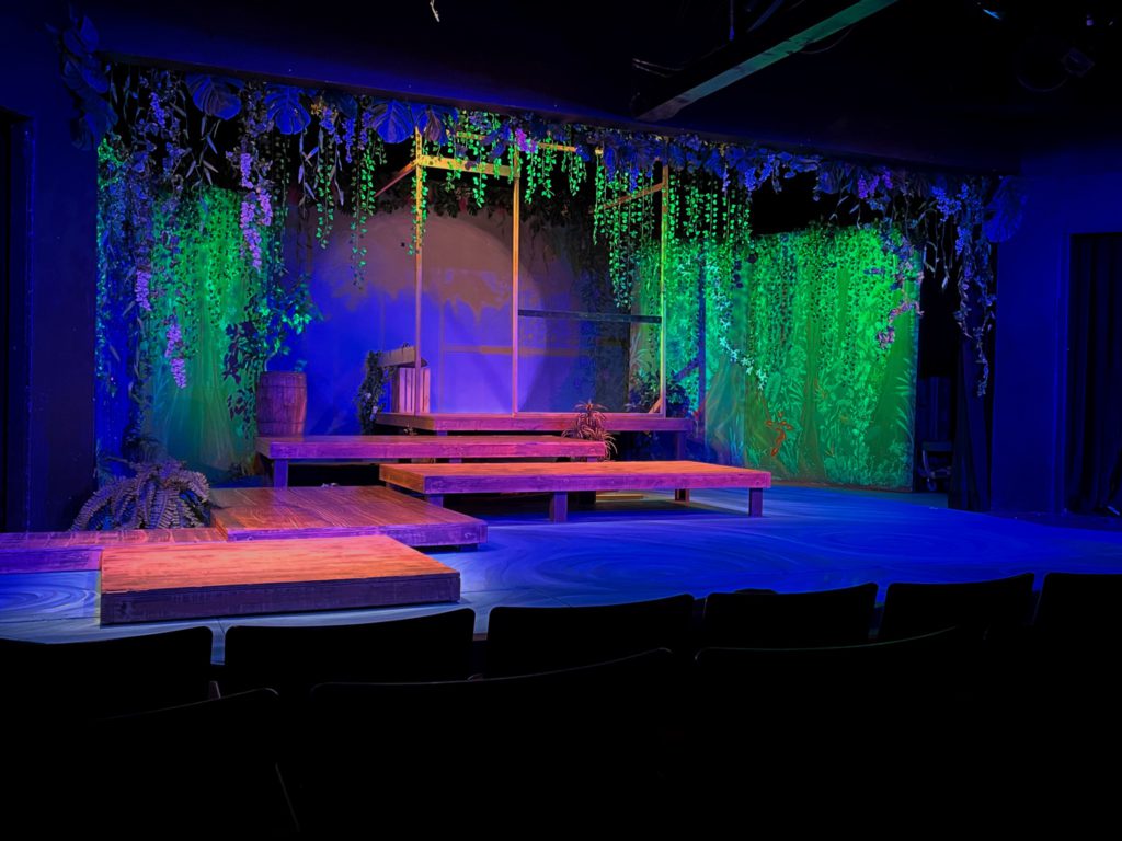 A forested backdrop with hanging flowers and vines framing a pier and swirling blue river invites the audience into the story. Photo Credit: Justin Kelley-Cahill